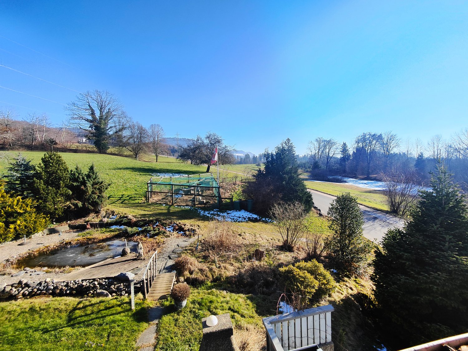 Aerial view of garden with trees and plants, a stream, and a winding road, surrounded by snow-covered ground and mountains in the distance.