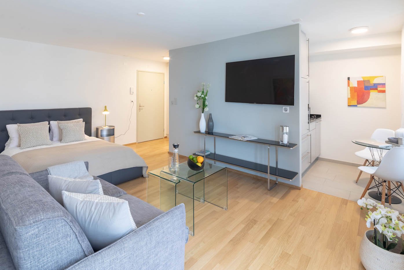 a modern and clean room with wooden floor, a double bed, a couch, TV on the wall, a side table with a lamp, a shelf with a bowl of fruits, and a dining area with chairs and a table.