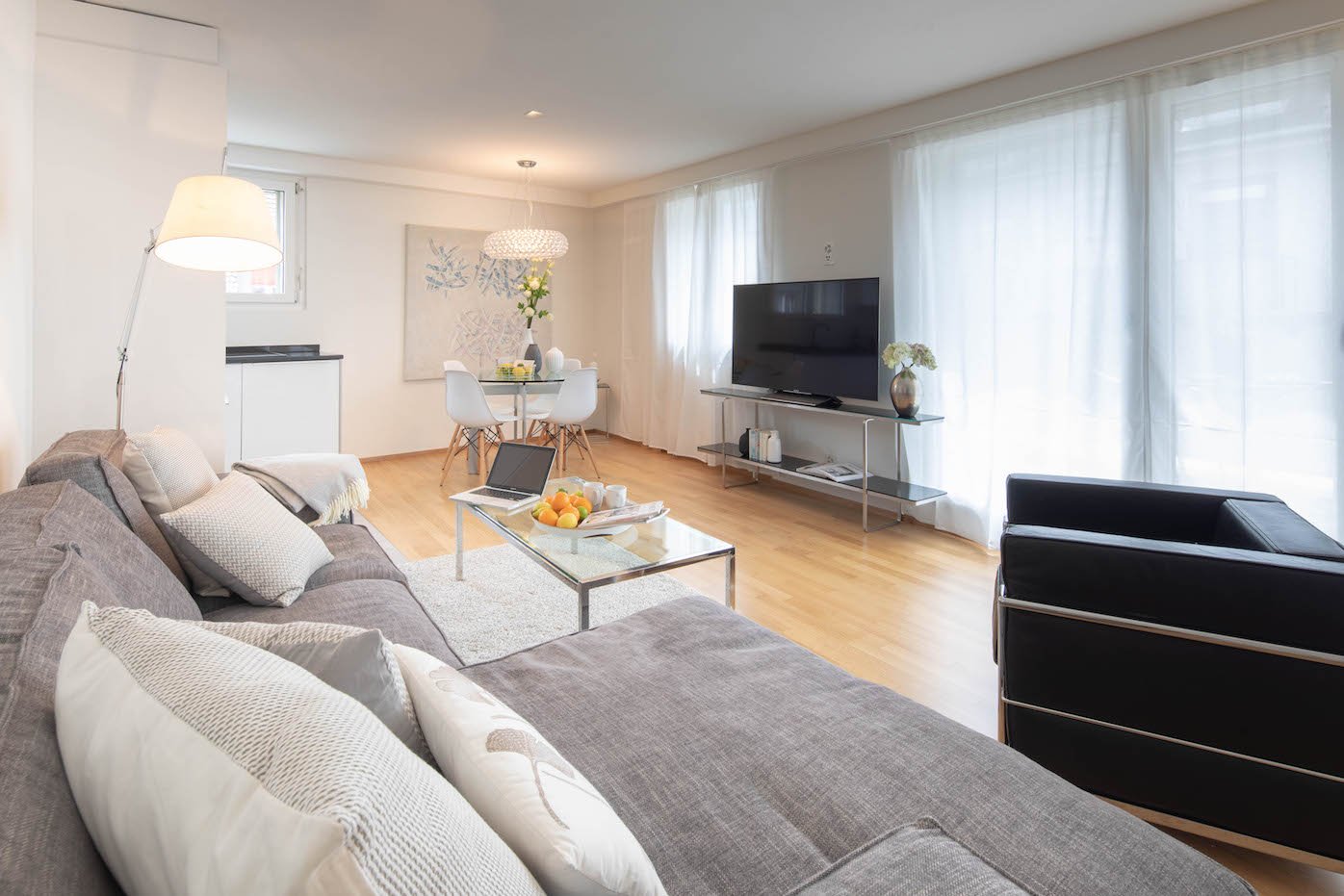 open space living room, couch, TV, coffee table, dining table, chairs, glass windows, floor lamp, white walls, wooden floor, vase, plant