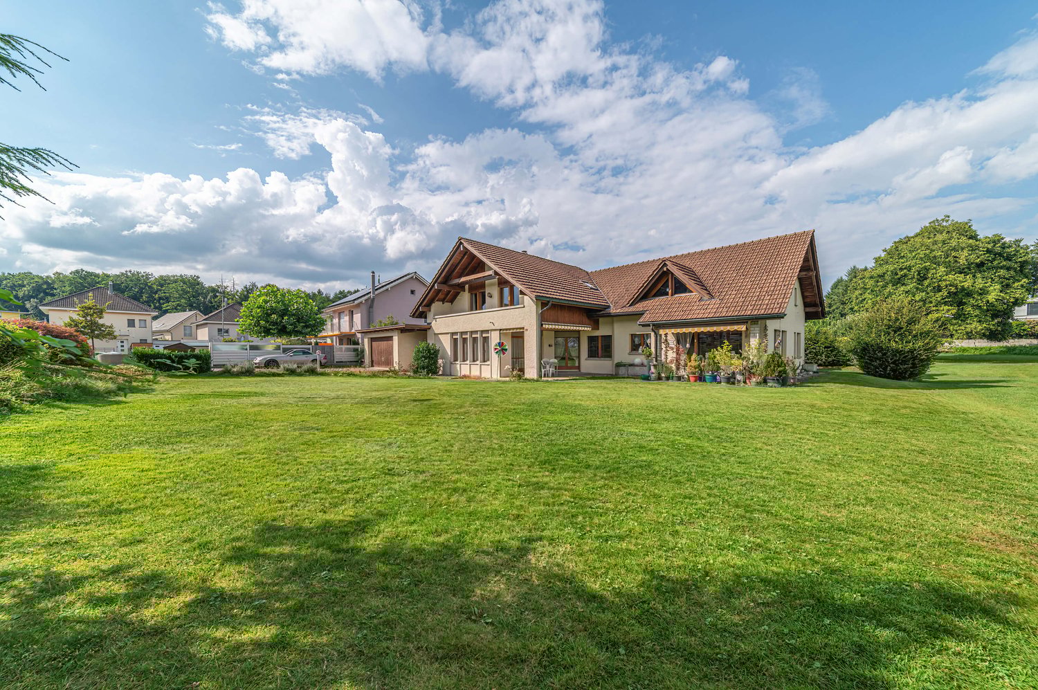 Detached house, brown roof, beige walls, windows with wooden frames, car in the driveway, large green lawn, several bushes and trees, cloudy sky