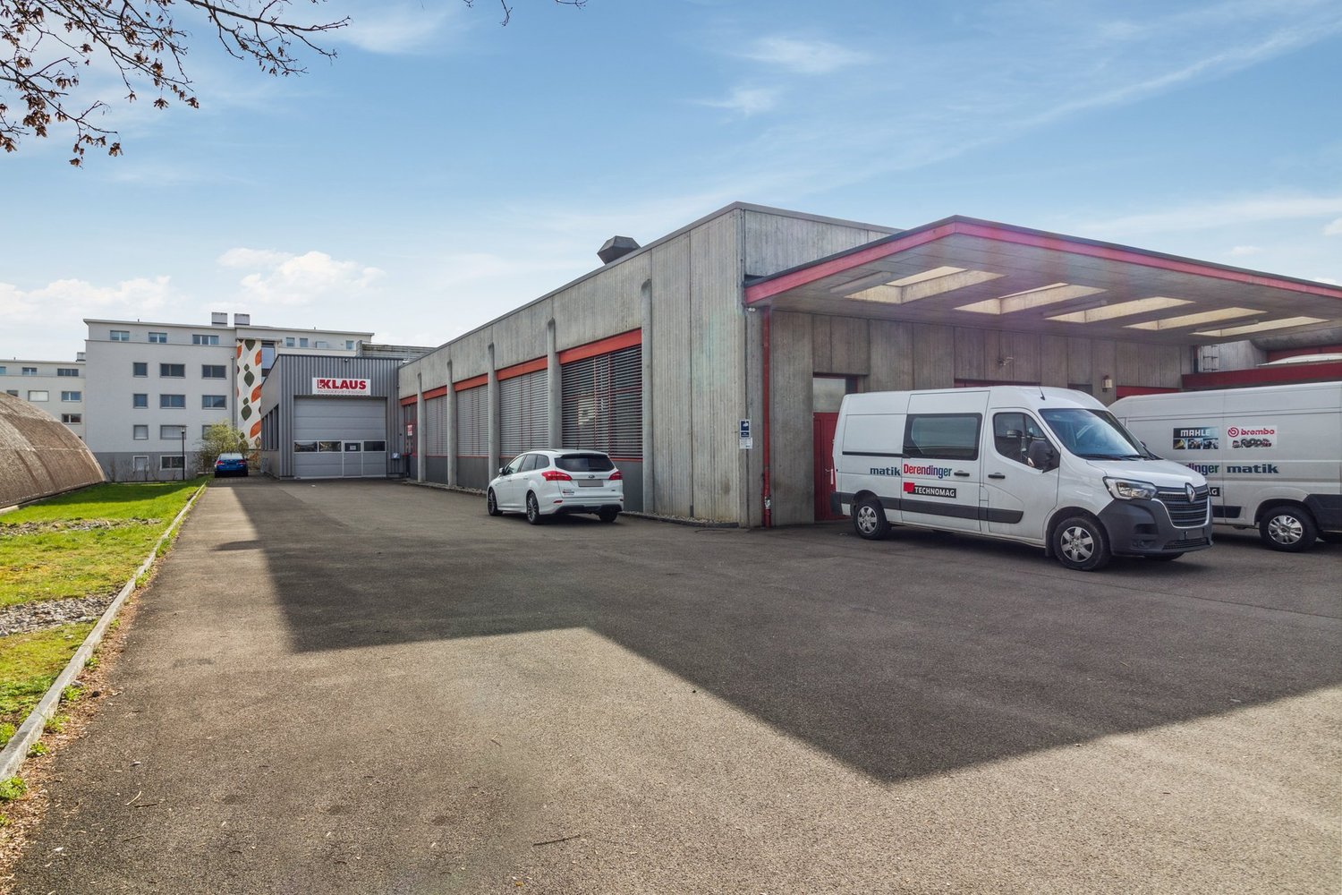 A large commercial building with a gray exterior and red accents. There are several vehicles parked in the paved lot in front of the building, including a white car and two delivery vans. The building appears to be a warehouse or industrial facility, with