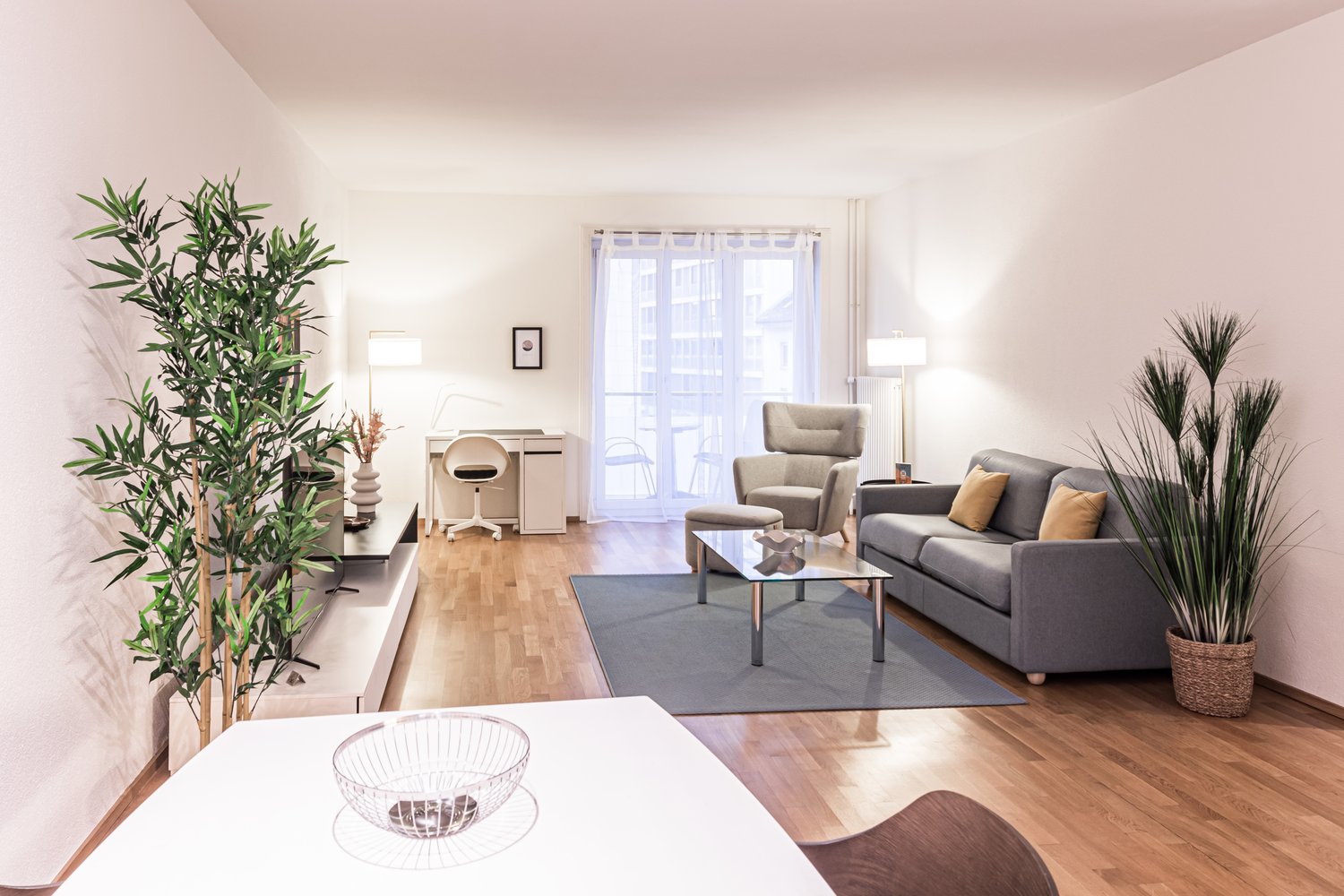 large windows, large gray couch, coffee table, glass table, armchair, desk, chair, two potted plants, wooden floor