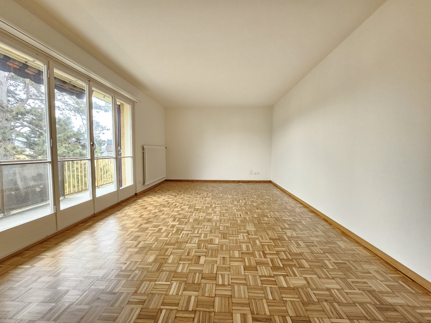 Spacious living room with large windows, hardwood parquet flooring, and white walls.