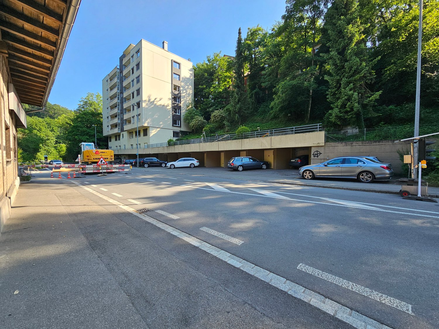 building with underground parking, cars parked in parking spaces, concrete wall, metal fence, plants and trees