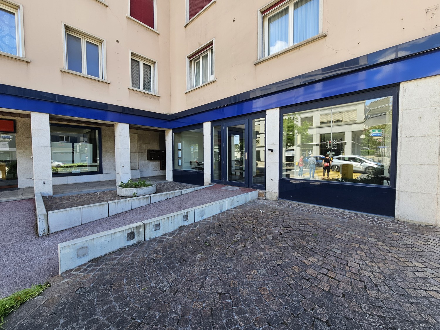 A multi-story commercial building with a blue-framed glass entrance and windows. The exterior walls are beige, and the ground floor has a paved entrance area with steps leading up to the entrance.