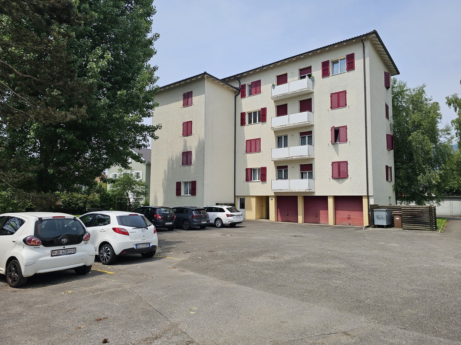 4-story apartment building with beige exterior and red shutters, parking lot in front with several cars parked