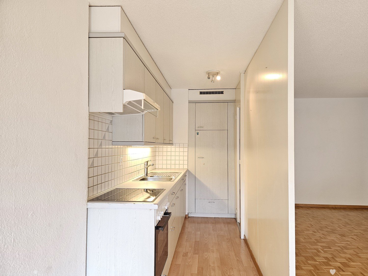 Small studio apartment with kitchenette, white cabinets, tiled backsplash, stove, dishwasher, and hardwood flooring. The apartment has a compact layout with an open floor plan.