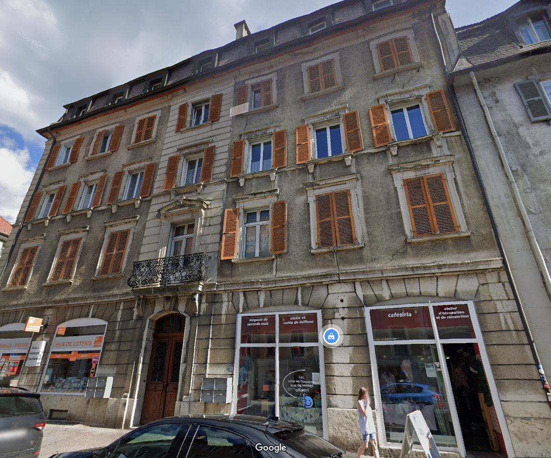 Multi-story building with a traditional European architectural style, featuring stone walls, wooden shutters, and a balcony. The building houses various commercial establishments, including a lottery shop, an art studio, and a cafeteria.