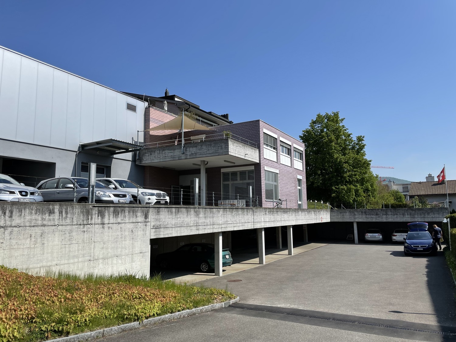A multi-story commercial or residential building with a parking area underneath. The building has a modern, angular design with a mix of materials including concrete, metal, and glass. There are several cars parked in the covered parking area below the bu