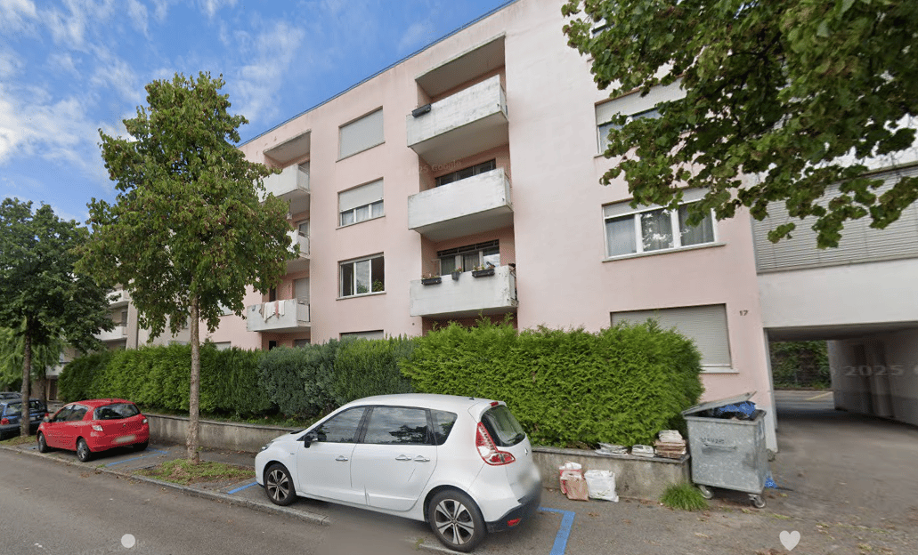 3-story apartment building with balconies, surrounded by trees and greenery, with parking spaces in front