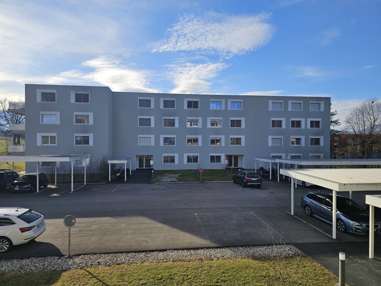 Multi-story apartment building with gray exterior, multiple windows, and a parking lot in the foreground with several cars parked.