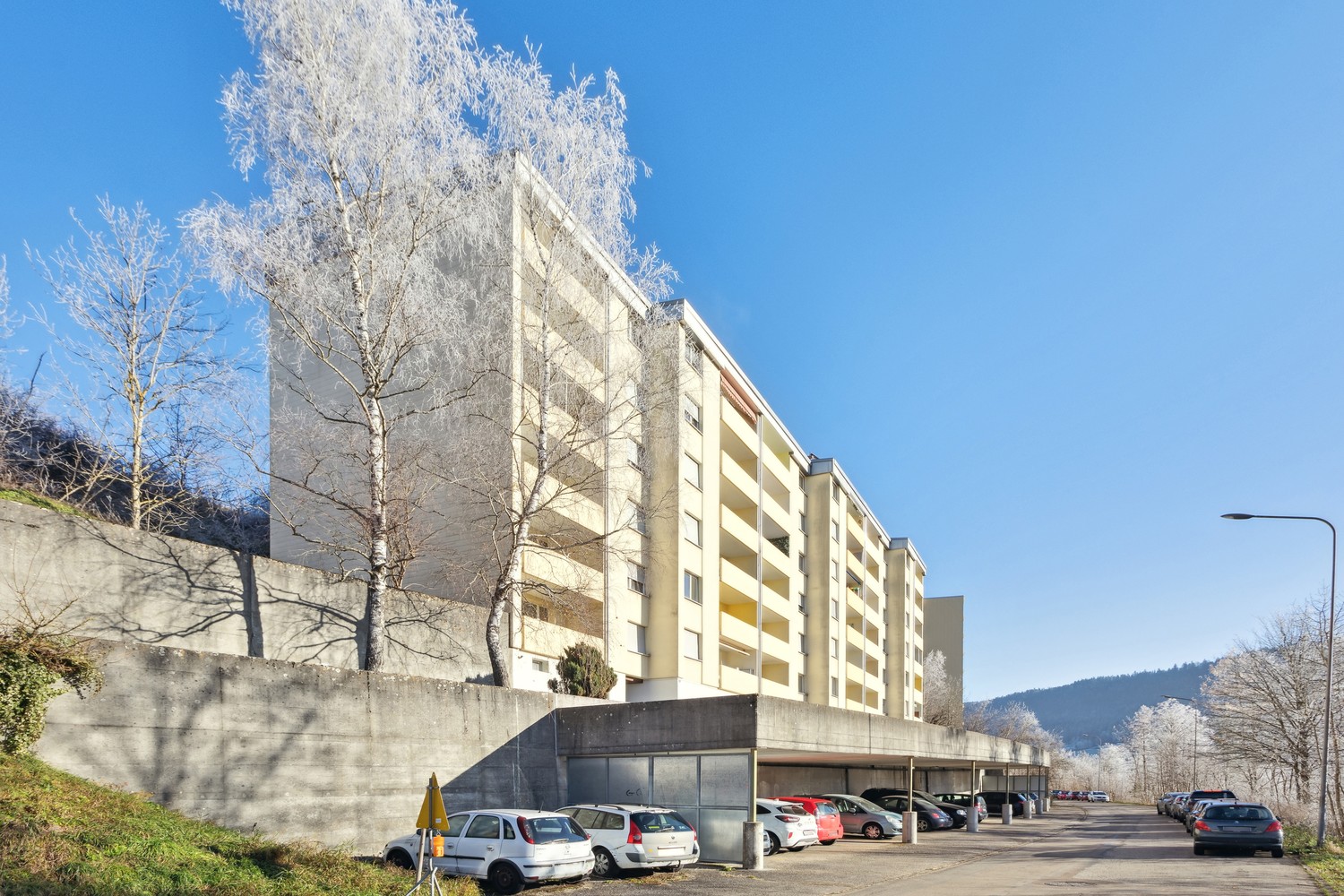 Multi-story apartment building with a parking lot in front, surrounded by trees covered in snow