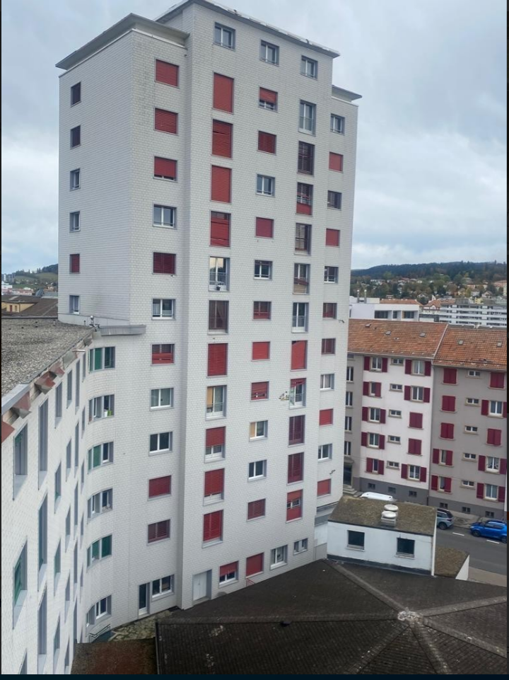 Multi-story apartment building with white exterior and red window frames, parking spaces visible in the foreground