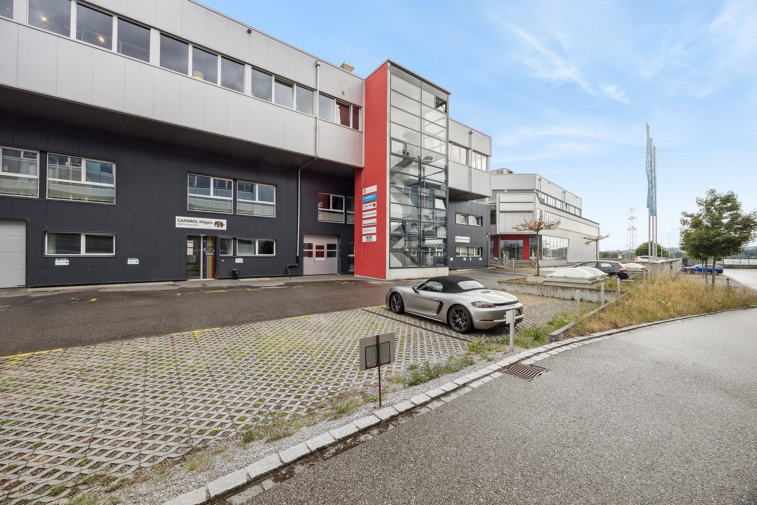 Multi-story commercial building with a modern, industrial design. The building has large windows, a red accent wall, and a parking area in front. There is a car parked in the lot.