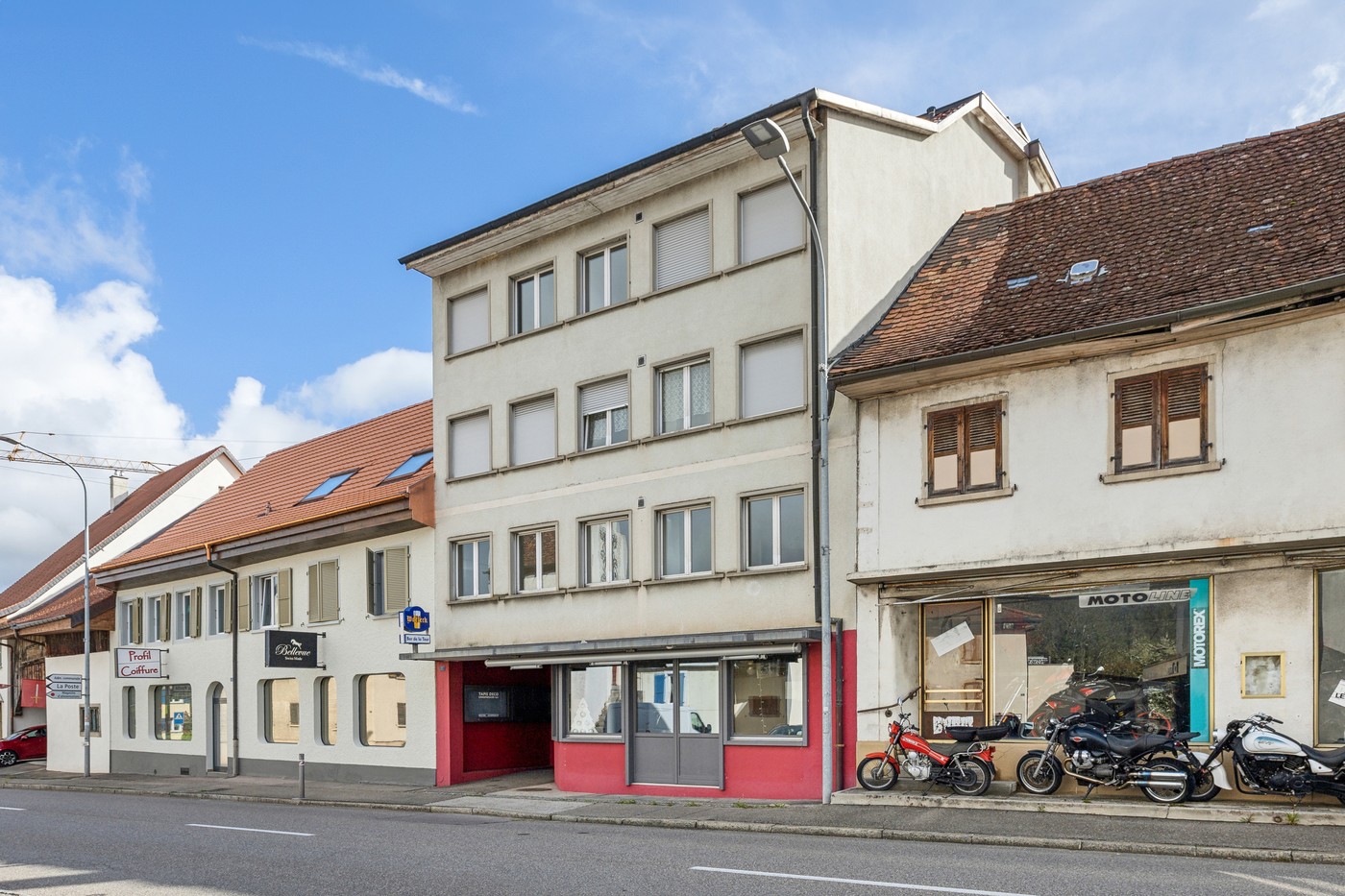 Multi-story building with a motorcycle shop on the ground floor, residential units above, red tile roof, and parked motorcycles in front of the building.