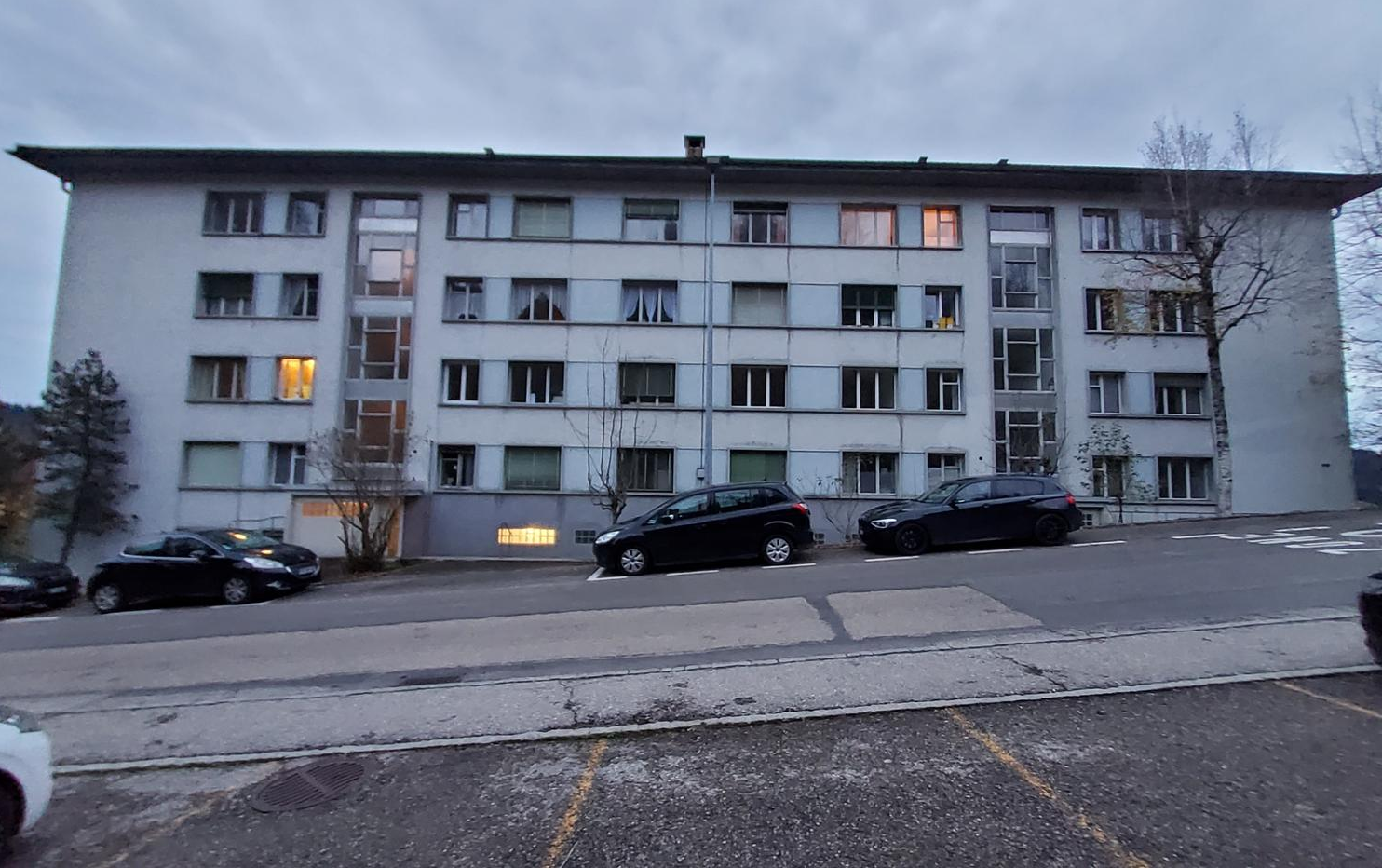 A multi-story apartment building with a gray exterior and several windows. There are parked cars in the foreground, indicating a parking area or lot nearby.