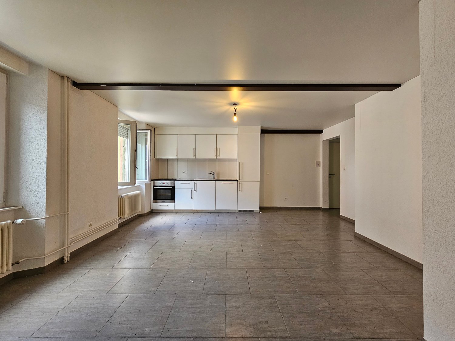 an empty room with white tiled floor, ceiling lamp, kitchen area with white cabinets, sink, and oven, radiators, and windows with bright sunlight