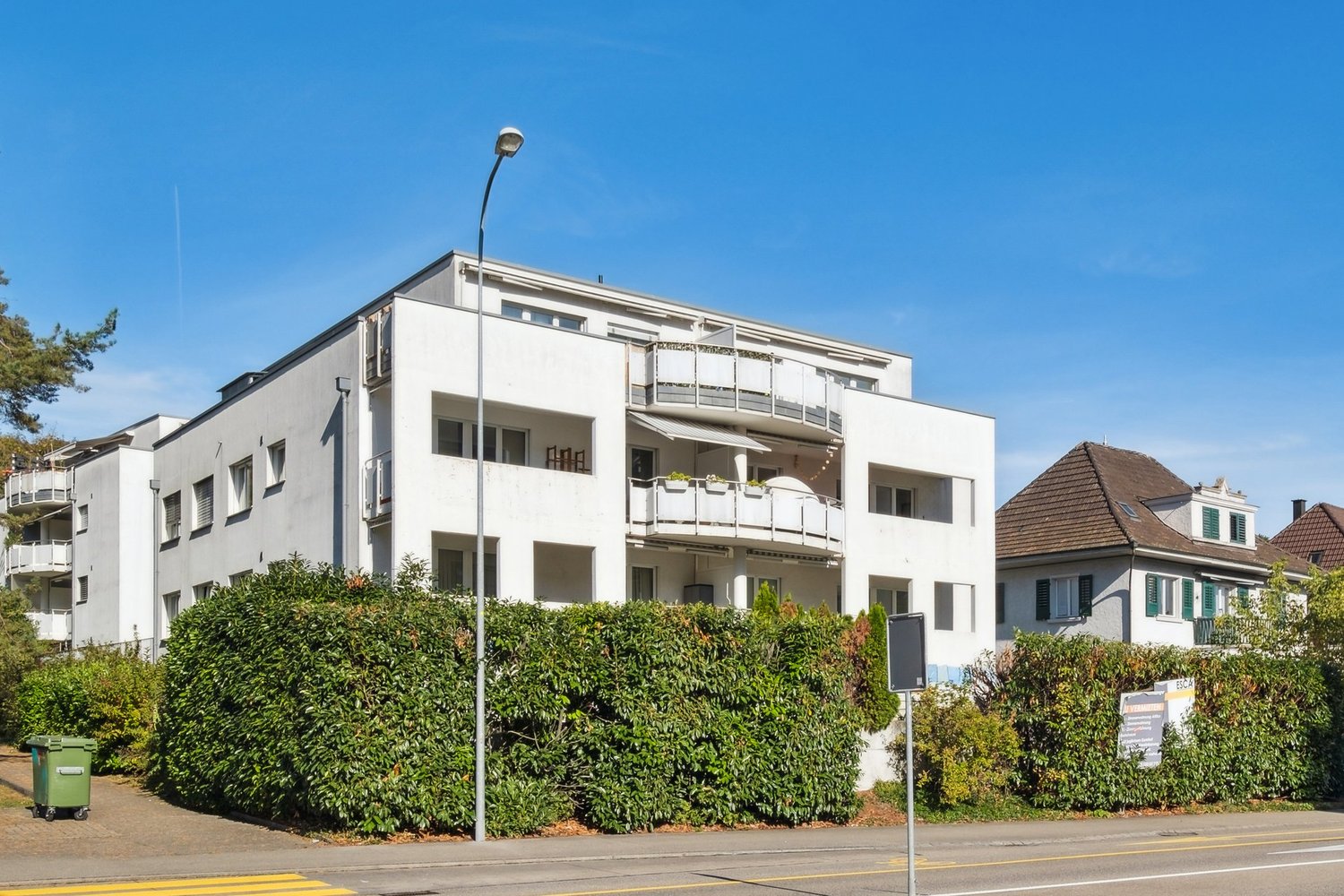 3-story apartment building, white, multiple balconies, hedges, trees