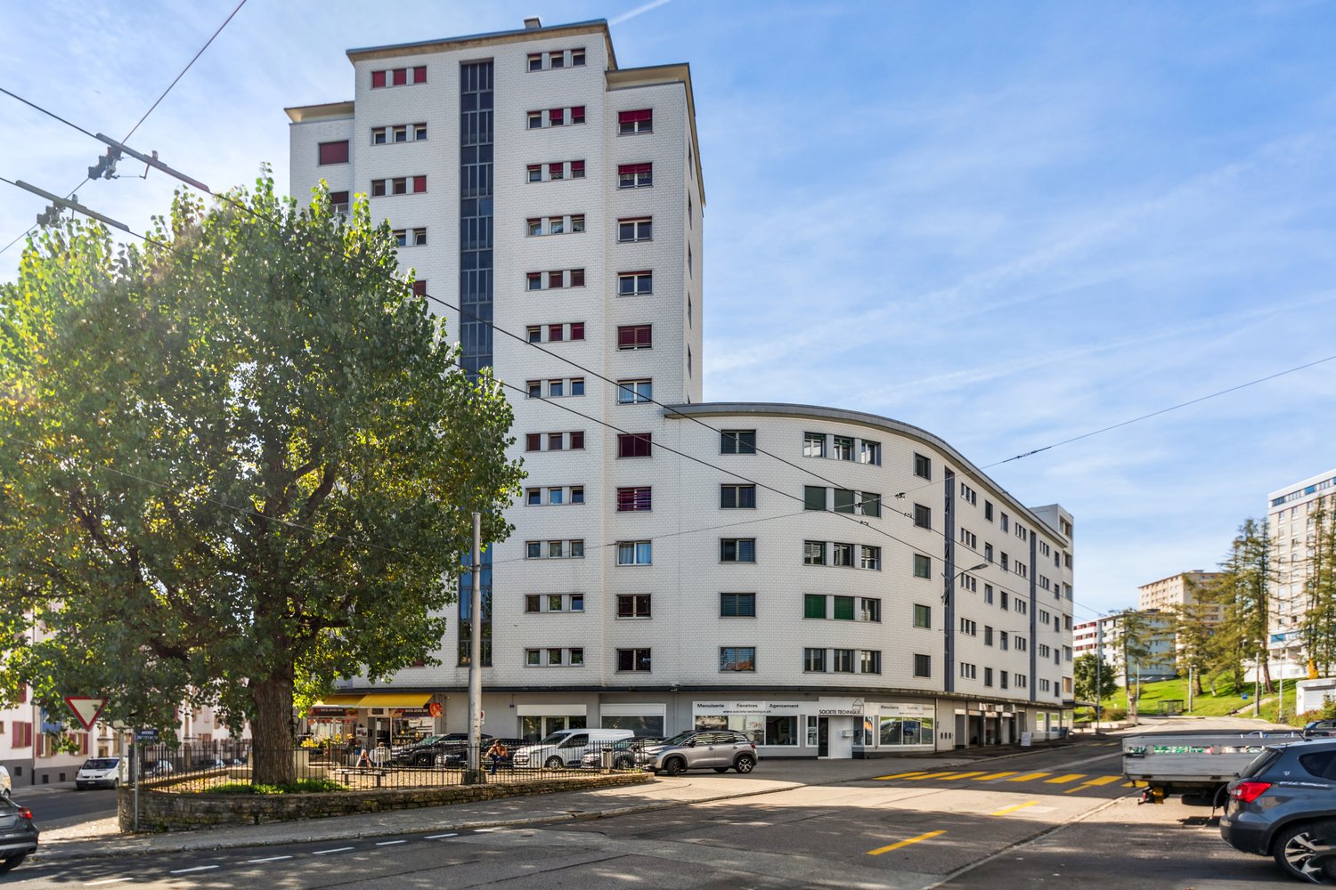 Modern multi-story building with white exterior and many windows, located at an intersection with a road and some parked cars.