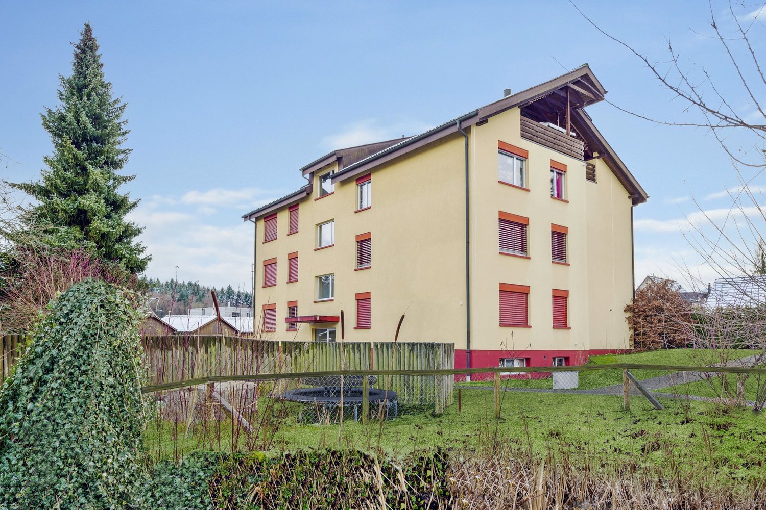 3-story yellow house with red trim, windows with red shutters, wooden fence surrounding the property, grassy field in front, small trampoline in the yard
