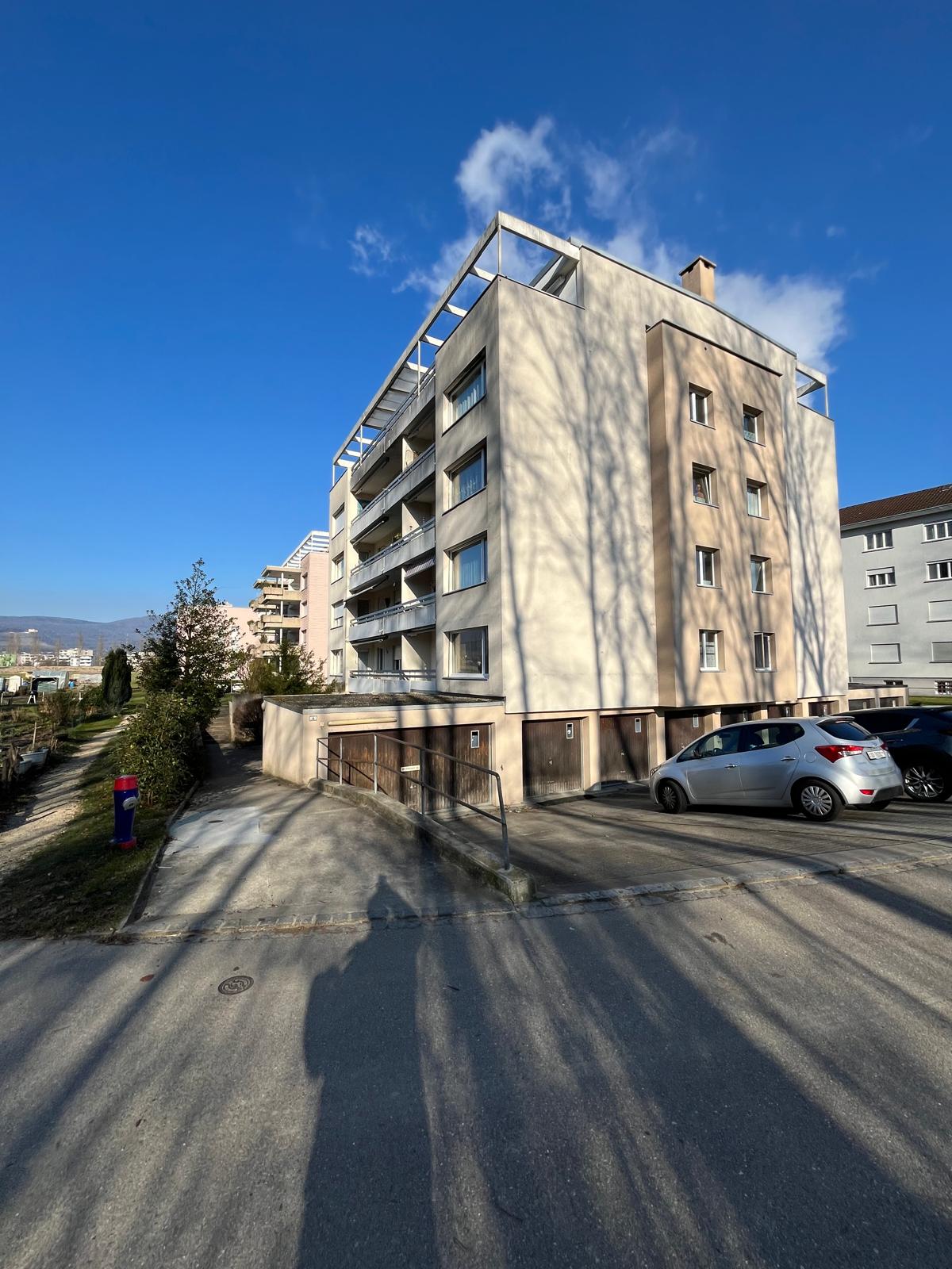 An apartment building with balconies, a car parking area in front, a building with a chimney on the top right, and some trees and greenery nearby.