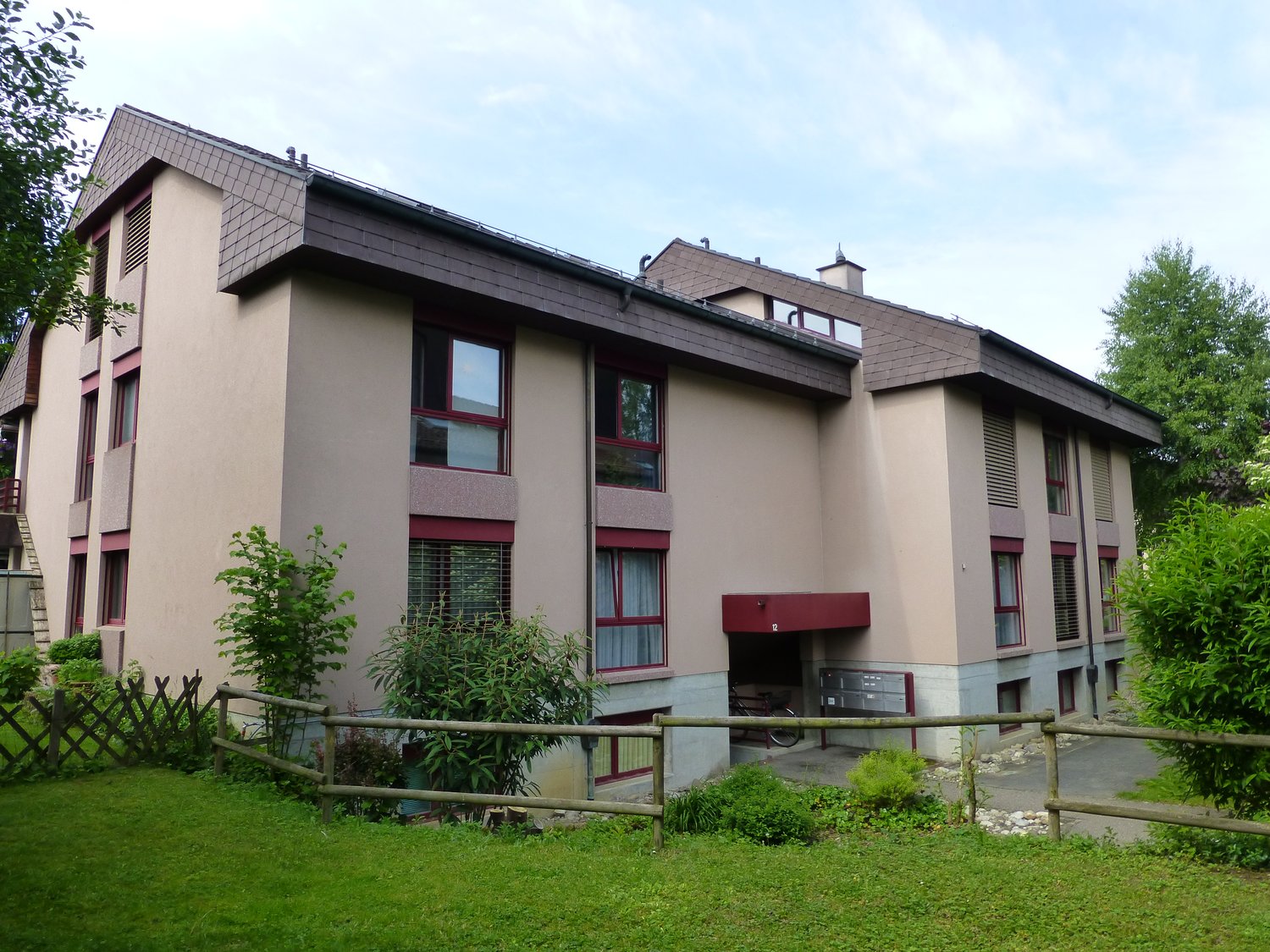 Multistory building, beige walls, red window frames, wooden fence, green lawn, staircases, red entrance