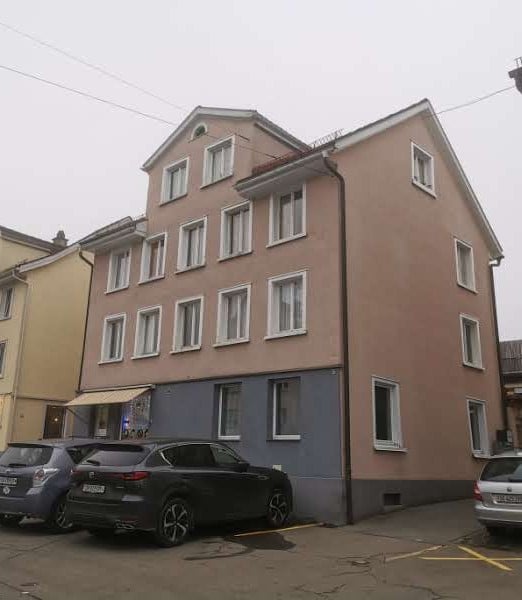 Three-story house with multiple windows, beige facade, awning, parking area