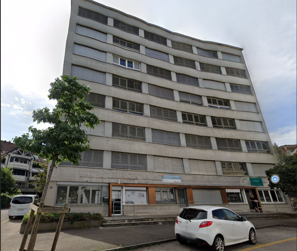 5 story office building, grey concrete, many windows, small tree in front, street parking