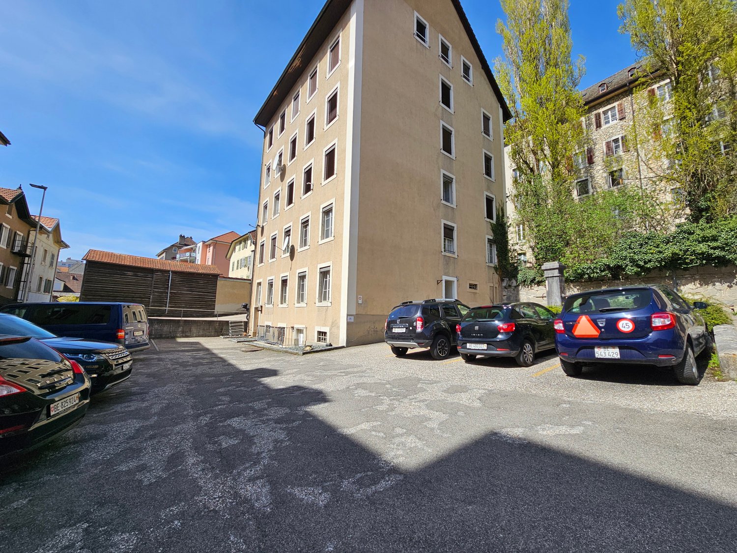 6 story building, multiple windows, parking area, 4 cars parked, red roof on nearby building, streetlight