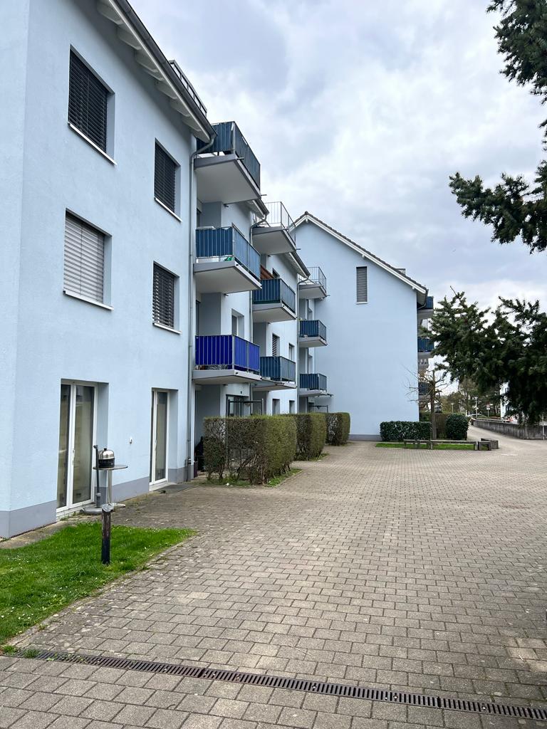 Blue apartment building with multiple balconies, brick paved driveway, hedges, trees