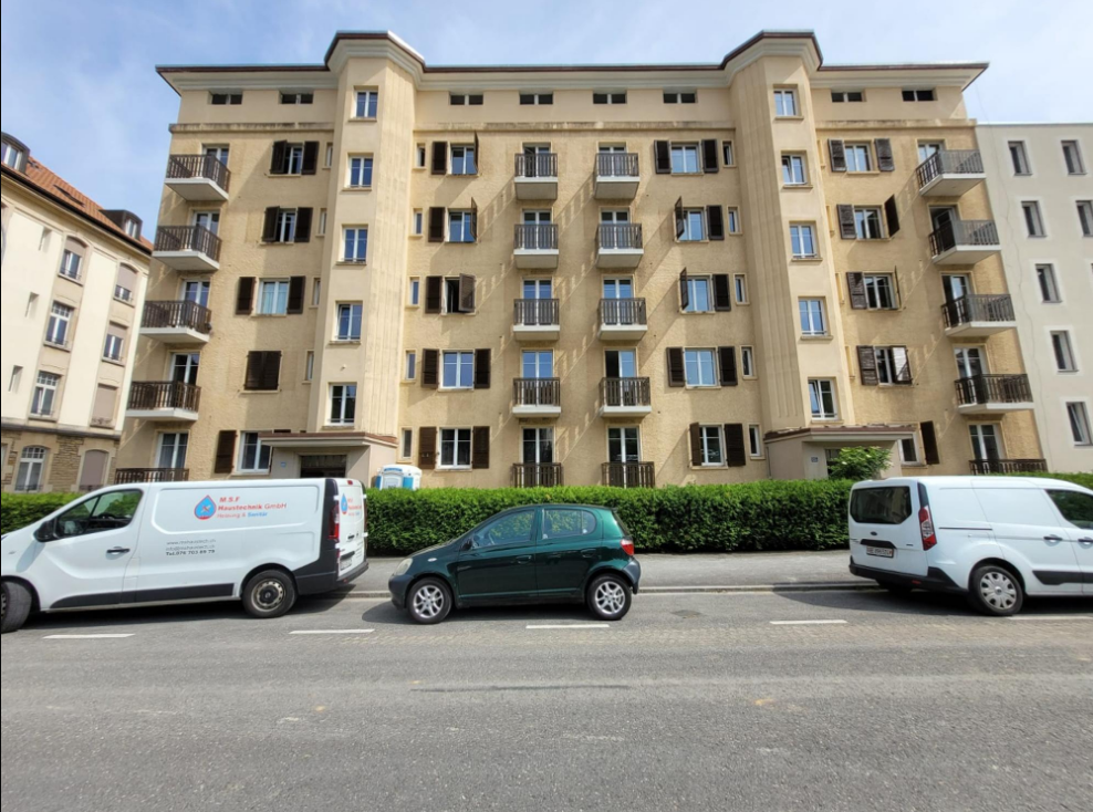 Multi-story apartment building with beige facade, balconies, and windows. Several parked cars visible in front of the building.