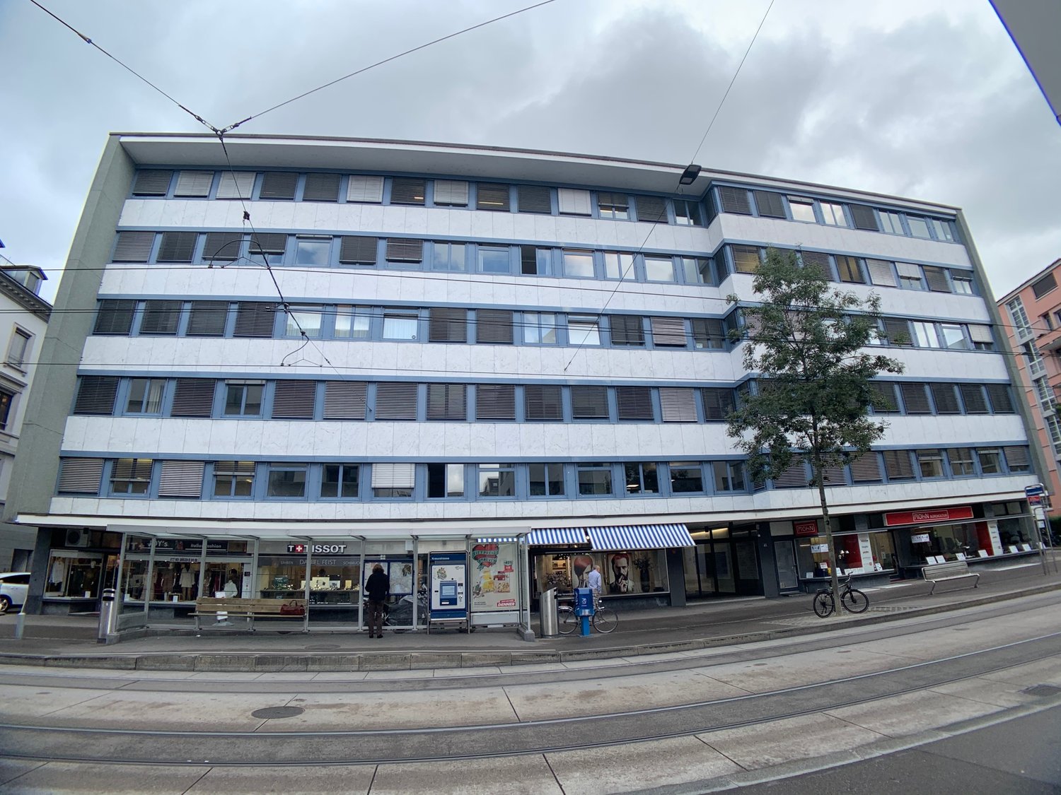 multi-story building with many windows, two stores on ground floor, bike parked in front, sidewalk