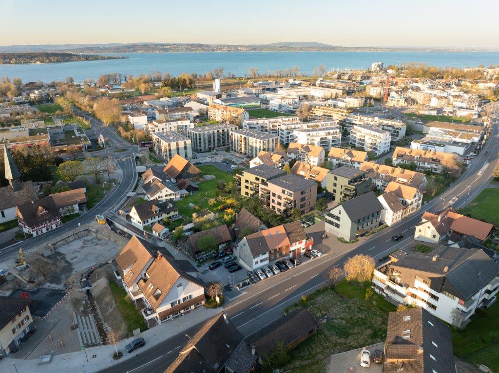Aerial view of a city with a mix of residential and commercial buildings, surrounded by a body of water and hills in the background. The city appears to have a mix of older and newer buildings, with some high-rise structures visible. The streets and roads