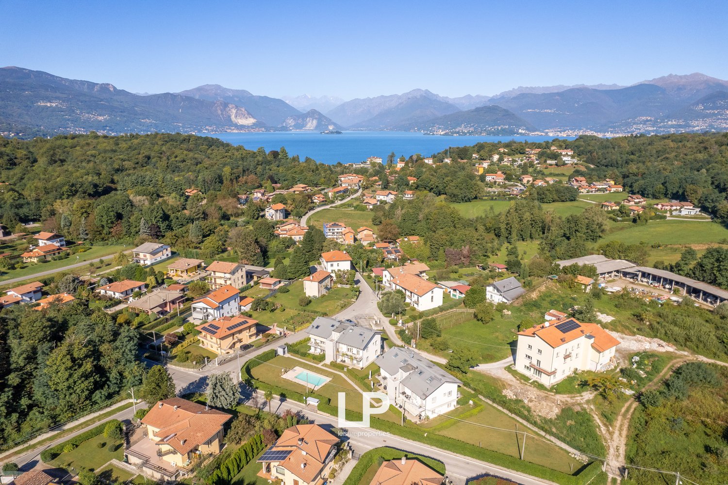 Numerous residential buildings, some with solar panels, surrounded by lush greenery, multiple houses with balconies, a swimming pool in front of a building, mountainous terrain, a large body of water in the background, and a clear blue sky.