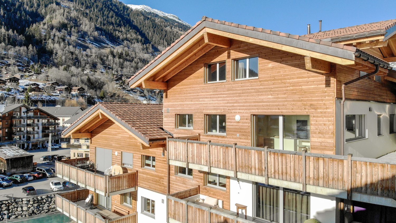 A multi-story wooden chalet-style building with a sloped roof and balconies overlooking a snowy mountain landscape. The building has large windows and a wooden exterior. There are other similar buildings and a village visible in the background.