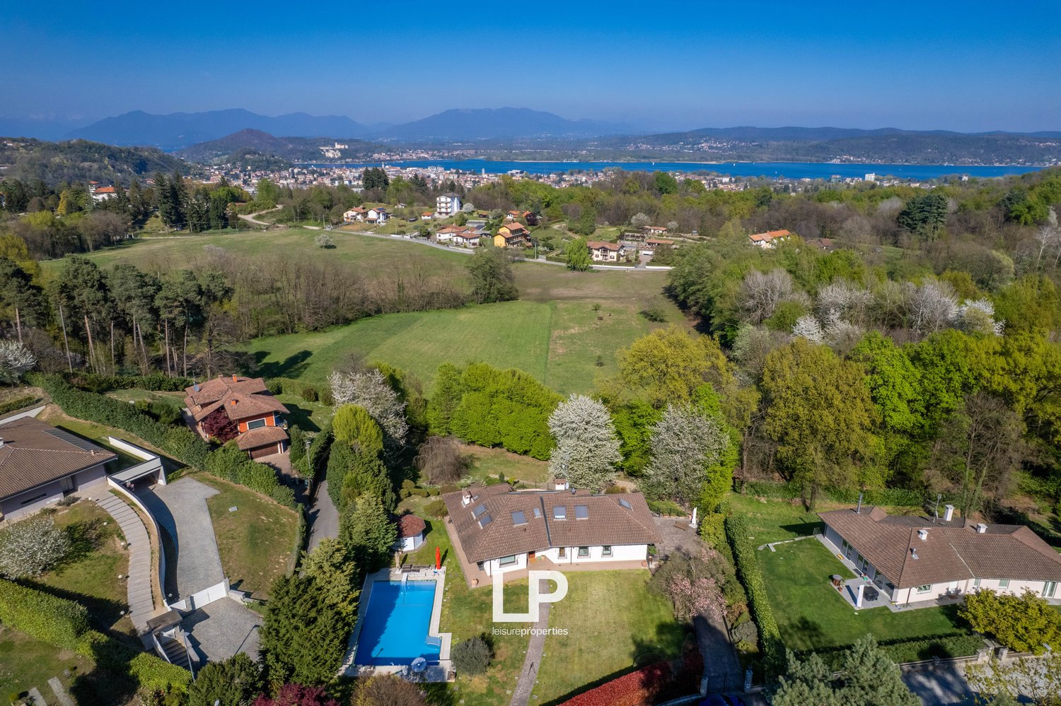 Aerial view of a large house with a swimming pool surrounded by lush greenery, with a scenic view of a lake and mountains in the background.