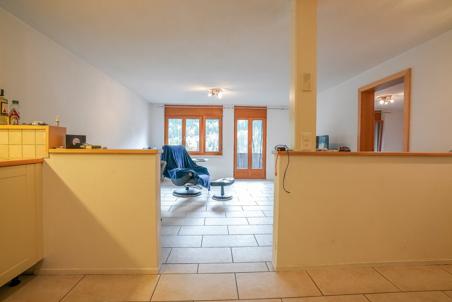 Spacious living room with large windows, wooden furniture, and a balcony or terrace visible through the glass doors.