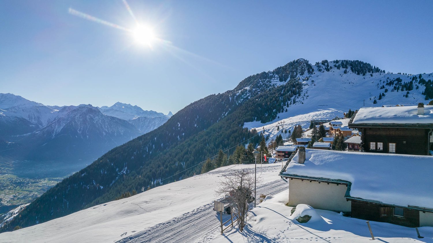 Snowy mountain landscape with a ski resort village in the foreground. The image shows snow-covered rooftops, ski slopes, and a scenic view of the surrounding mountains under a bright, sunny sky.