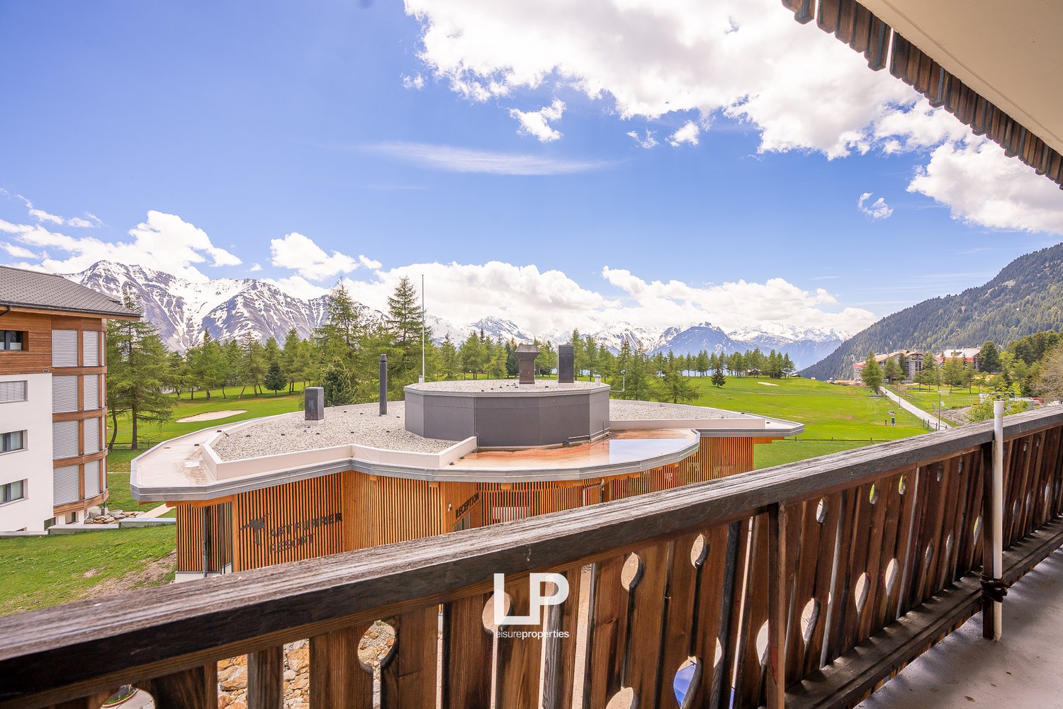 Balcony overlooking a scenic mountain landscape with snow-capped peaks, green meadows, and a fountain or water feature in the foreground. The balcony has a wooden railing and the apartment building appears to be in a resort or alpine setting.