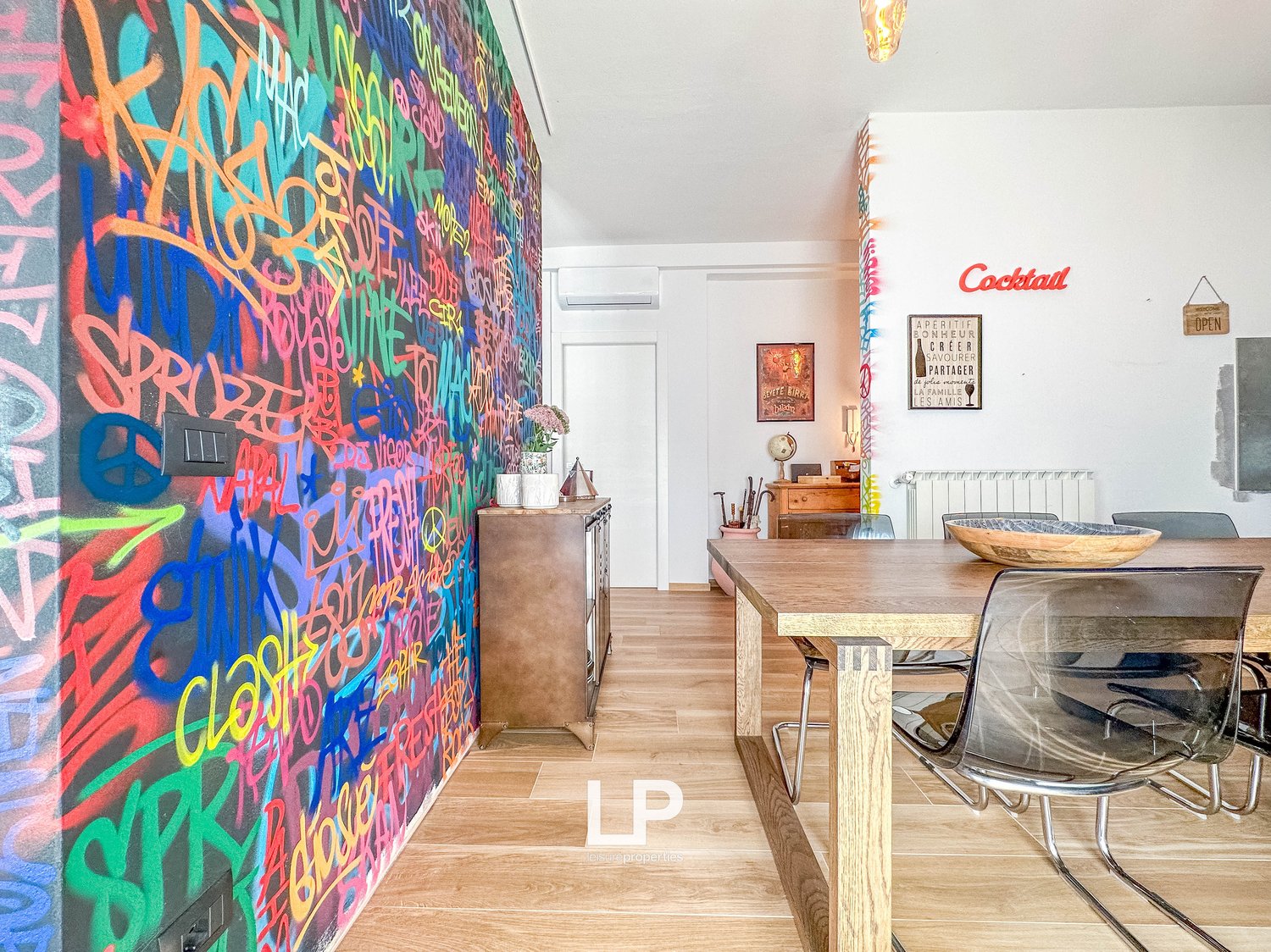 Colorful graffiti-style mural wall, wooden dining table with chairs, shelving unit, and a fireplace visible in the room.