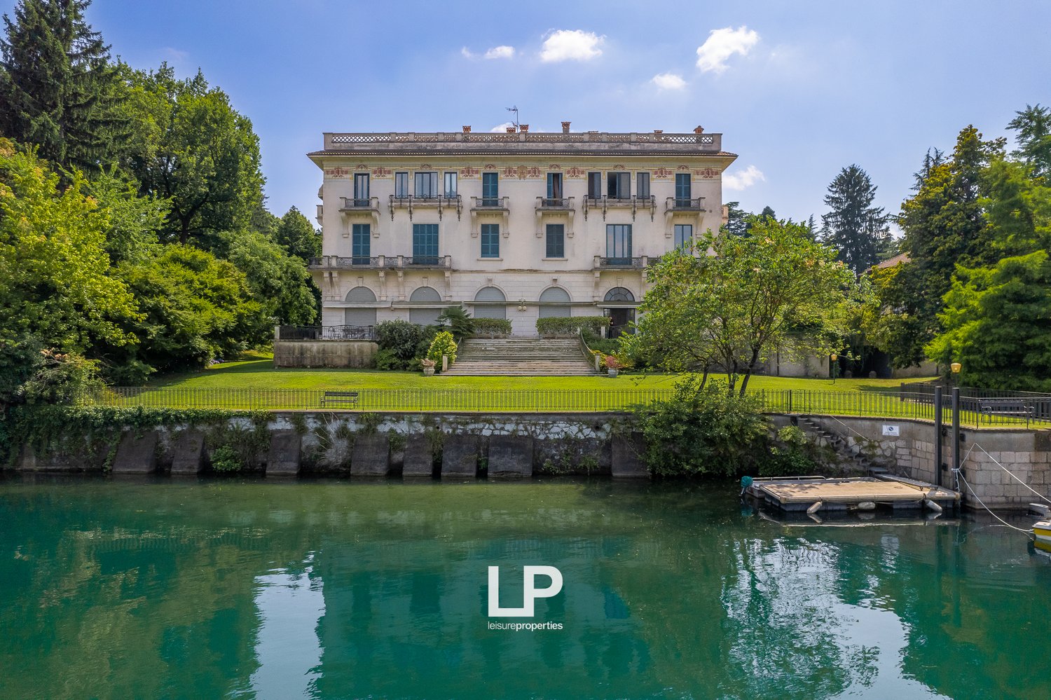 A large, multi-story historic building with a classical architectural style, surrounded by lush greenery and a body of water. The building has balconies, arched windows, and a decorative roofline.