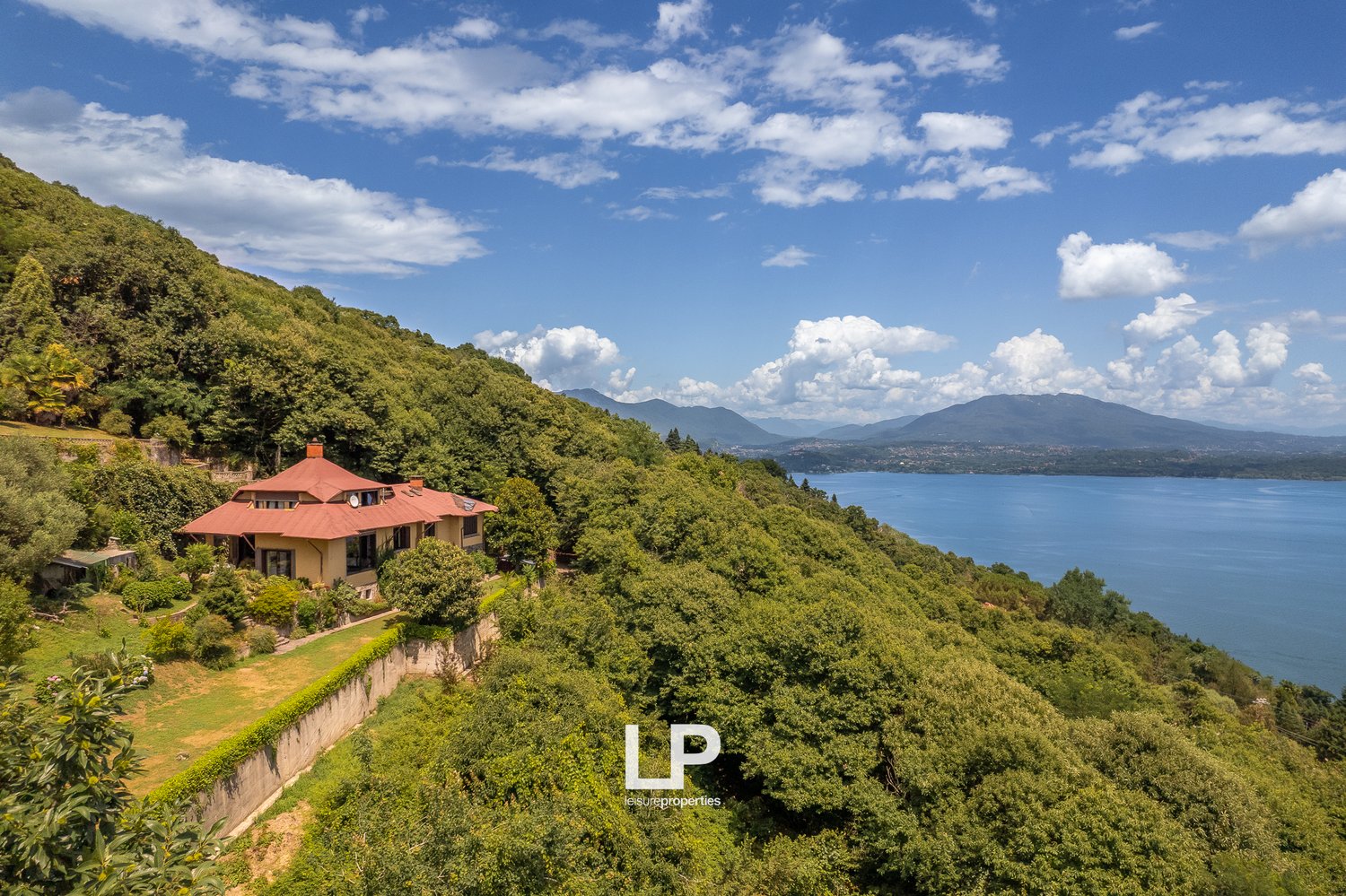 A large, two-story house with a red roof situated on a hillside overlooking a lake and mountains. The house is surrounded by lush, green vegetation and trees. The landscape features a scenic view of the lake and mountains in the distance.
