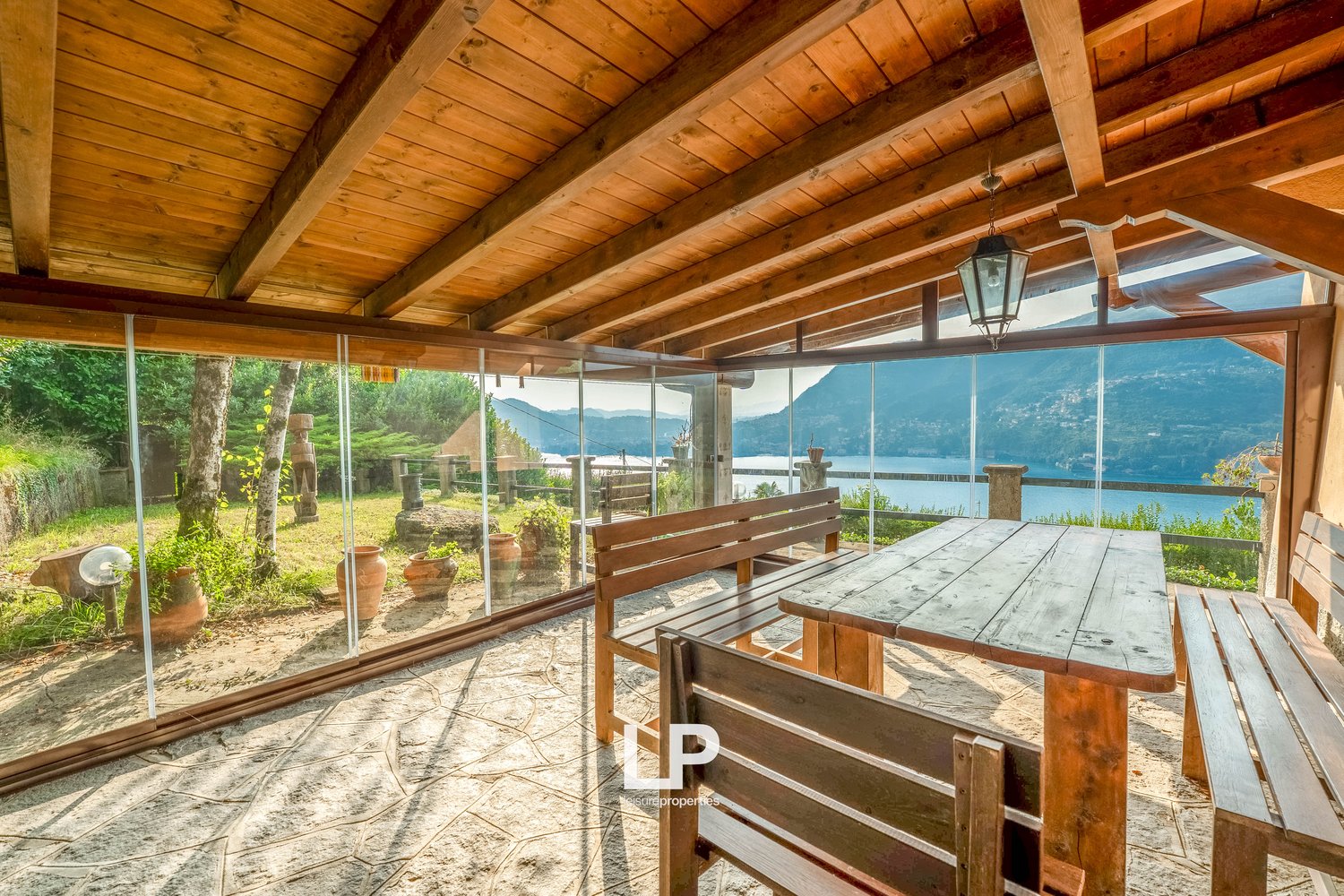 Enclosed terrace, wooden benches and table, glass walls, hanging lamp, view of mountains and water, decorative potted plants on ground