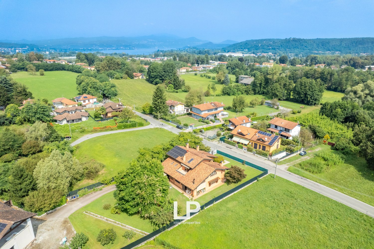 Detached house, red roofs, green landscape, visible solar panels, large green fields, mountain view in the distance, swimming pool on the side