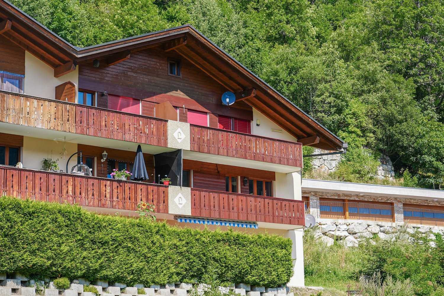 Apartment building, multiple wooden balconies, red shutters, hedge, grassy hillside, stone retaining wall