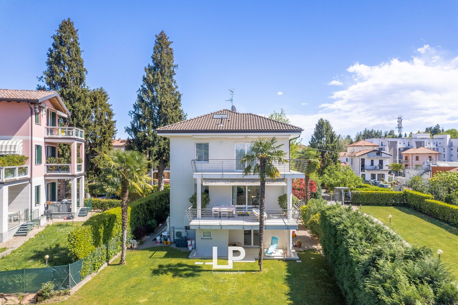 Two-story house with a tiled roof, surrounded by lush greenery and palm trees. The house has balconies, a garage, and parking spaces. The property is situated in a residential area with other houses and buildings visible in the background.