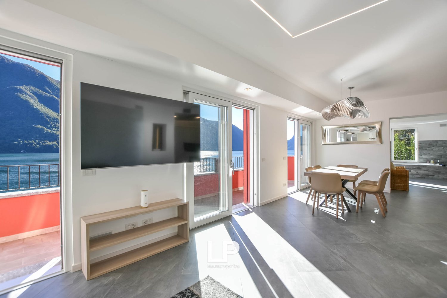 modern living room, gray floor, white walls, large window, flat screen TV mounted on wall, wooden table and chairs, dining area with open balcony, view of mountains and lake, decorative lighting