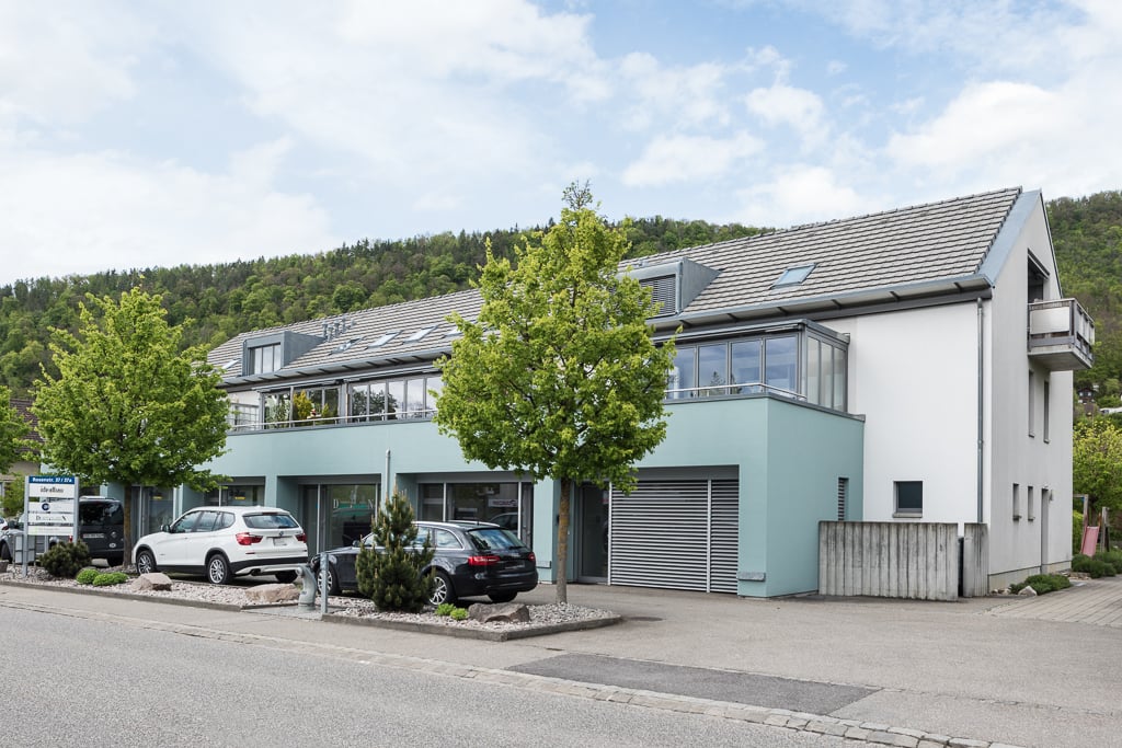 2-story commercial building with a gray roof, white walls, and large windows. There are several cars parked in the front of the building. The building is surrounded by trees and greenery.