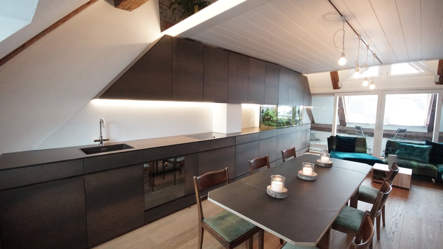 The image shows a modern, open-concept kitchen and dining area with a vaulted ceiling. The kitchen features dark, minimalist cabinets and a sleek countertop with a built-in sink. A dishwasher is visible in the kitchen. The dining area has a large wooden t