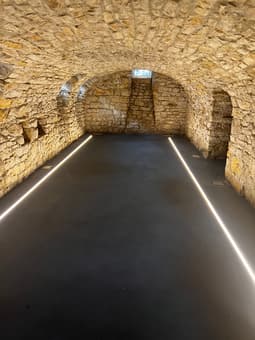 A long, dark tunnel with a paved floor and stone walls. The tunnel appears to be underground or within a building, with arched ceilings and lighting fixtures visible.